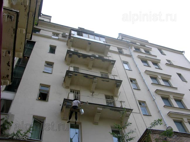  промышленный альпинист стал готовить балконы к покраске, сейчас он грунтует валиком балконные плиты
