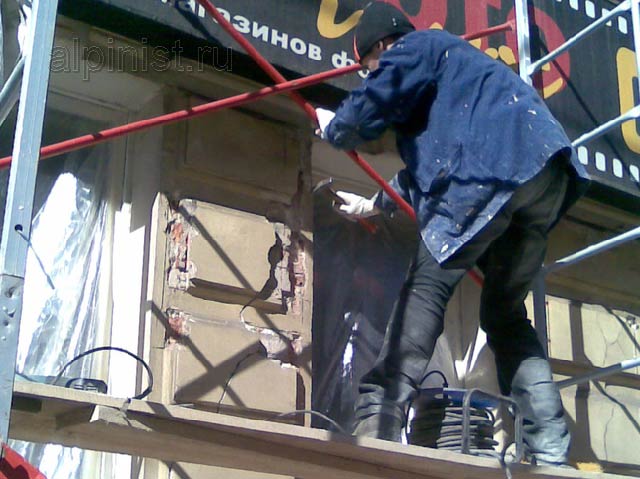 В данный момент мастер отбивает молотком бухтящую штукатурку в части фасада между окнами, где были большие разрушения.