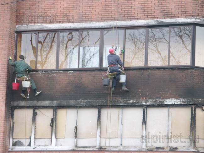альпинисты моют окна балкона и очищают кирпичный фасад здания средством для очисти фасада от копоти